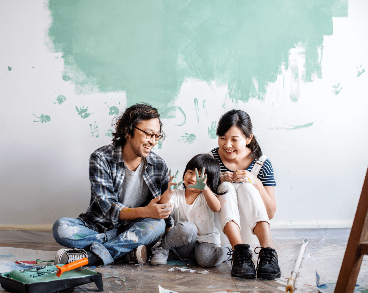 Man kissing baby's cheek. Couple in front of a painted wall in mid-progress smiling with their child, who’s hands are covered in paint