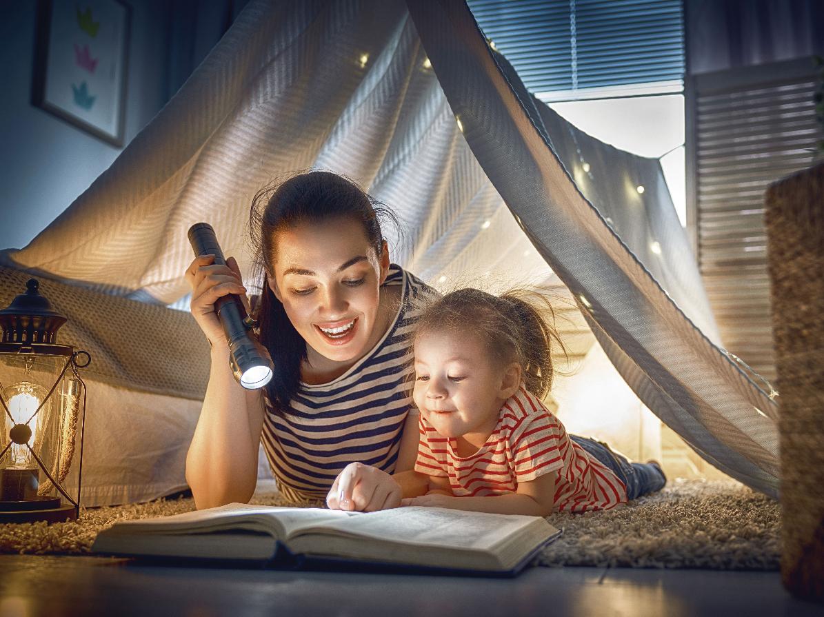 A woman and a child using a flashlight to read a book under a blanket.